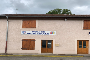 Polizeigebäude mit einem Schild, Fenstern, Türen, einer Namensplatte, einem Rohr, einem Baum und einem bewölkten Himmel im Hintergrund.