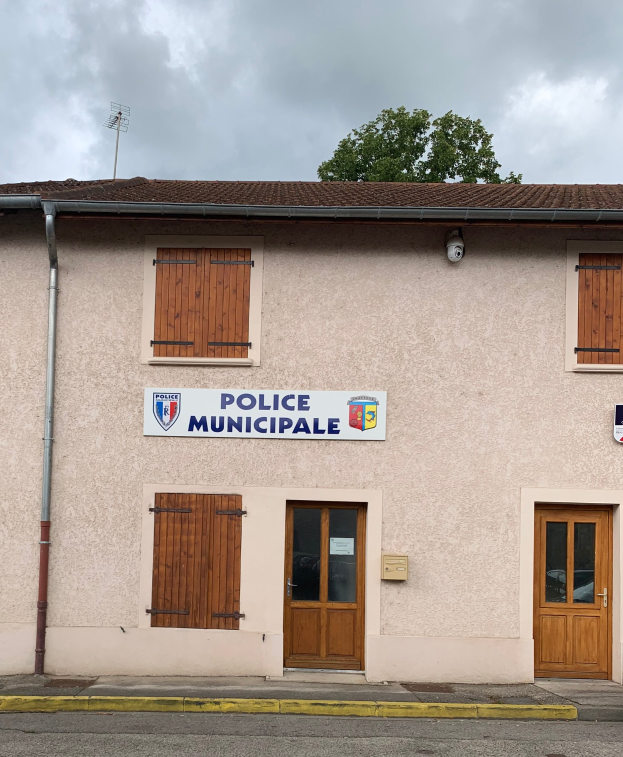 Polizeigebäude mit einem Schild, Fenstern, Türen, einer Namensplatte, einem Rohr, einem Baum und einem bewölkten Himmel im Hintergrund.