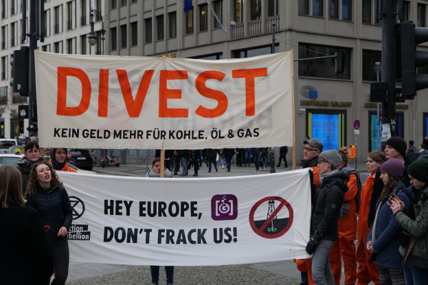 Gruppe von Menschen mit einem "Divest Hey Europe, Don't Frack Us"-Schild auf einer Straße mit Gebäuden, Fahrzeugen und Bäumen im Hintergrund unter einem klaren blauen Himmel.