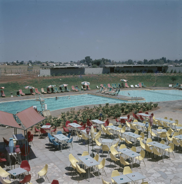 Ein Freibad mit Tischen, Stühlen und Sonnenschirmen umgeben von Pflanzen, einem Zaun, einer Wand, Bäumen und Gebäuden unter einem bewölktem Himmel.