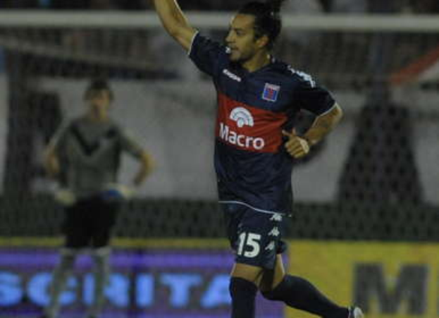 Ein Fußballspieler feiert nach einem Tor auf dem Boden, mit jubelnden Zuschauern und einer Person im Hintergrund, alles vor einem leicht unscharfen Hintergrund.