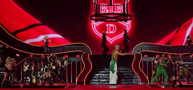 Eine Gruppe von Menschen auf einer Bühne, einige halten Musikinstrumente, mit Stufen, die zur Bühne führen und einem Schild im Hintergrund mit der Aufschrift "Rihanna - Live in der O2 Arena, London".