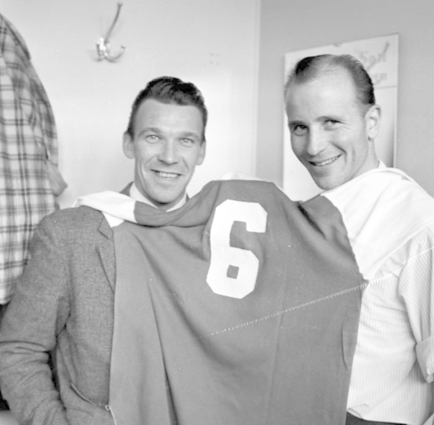 Zwei Männer mit einem Fußballtrikot in den Händen, die vor einem Schwarz-Weiß-Foto mit einer Tafel und einem Bügel an der Wand stehen.
