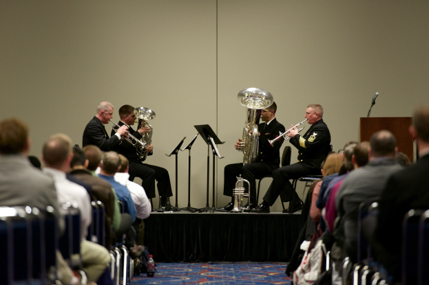 Männergruppe spielt Blechblasinstrumente auf einer Bühne mit Notenständern, einem Podium mit Mikrofon und einem Publikum auf Stühlen sitzend.