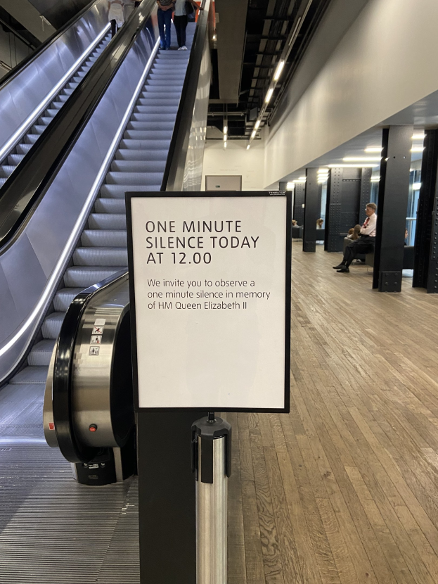 Eine Rolltreppe im Flughafen mit einem Schild, auf dem "Eine Minute Stille heute" steht, sowie ein paar Menschen darauf und Lichter an der Decke im Hintergrund.