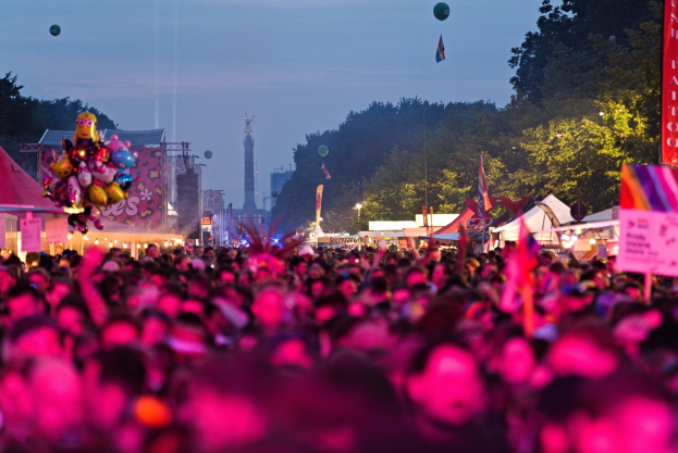 Eine große Menschenmenge bei einem nächtlichen Musikfestival mit farbigen Lichtern, Ballons, Zelten, Bannern, Fahnen, Bäumen, einem Turm und einem sternenklaren Himmel im Hintergrund.