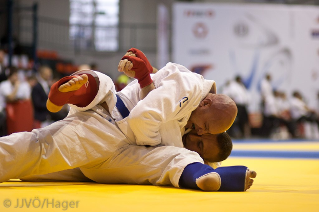 Zwei Judo-Kämpfer im Match, mit Zuschauern und Innenraum-Elementen im Hintergrund und Text unten.