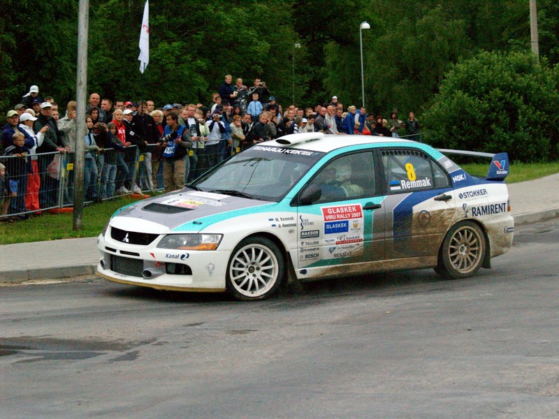 Ein Rallyeauto mit Text und Logos fährt an einer Menge vorbei, die Kameras hält, mit Geländern, Fahnenmasten und Bäumen im Hintergrund.
