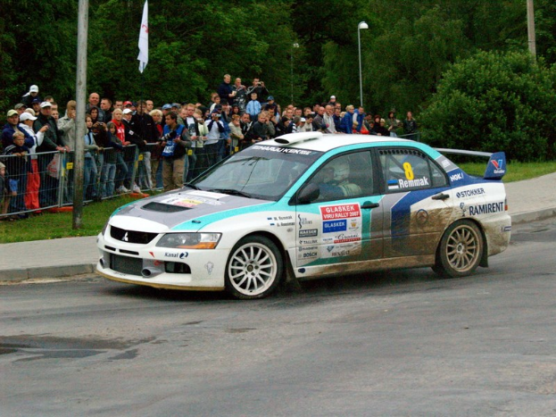 Ein Rallyeauto mit Text und Logos fährt an einer Menge vorbei, die Kameras hält, mit Geländern, Fahnenmasten und Bäumen im Hintergrund.