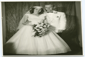 Ein Schwarz-Weiß-Foto einer Braut in einem weißen Kleid, die einen Strauß hält und neben ihr ein strahlender Bräutigam steht, mit einem Vorhang im Hintergrund.