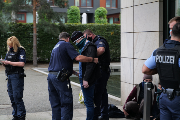 Gruppe von Polizeibeamten vor einem Gebäude mit einer Frau, die ein Buch und einen Stift hält, mehreren Menschen in der Nähe und Bäumen und Gebäuden im Hintergrund.