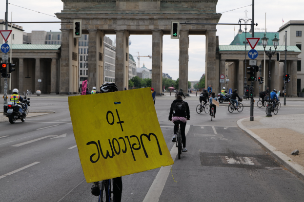 Eine Gruppe von Radfahrern fährt an der Brandenburgertor in Berlin vorbei, einer hält ein gelbes Schild, mit Laternenpfählen, Verkehrszeichen, Gebäuden und Bäumen im Hintergrund und einem klaren blauen Himmel.