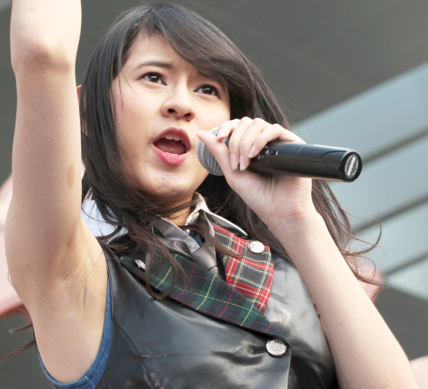 Frau singt leidenschaftlich in ein Mikrofon auf der Bühne, trägt ein weißes Hemd und einen Pferdeschwanz, hat die Augen geschlossen und einen konzentrierten Gesichtsausdruck, vor einem Gebäudehintergrund.