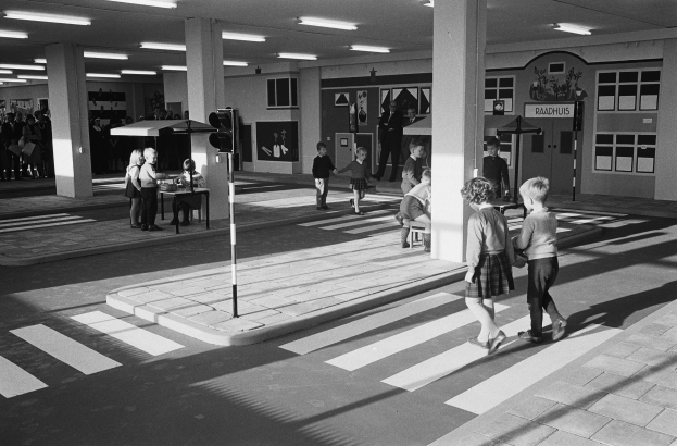 Schwarz-weißes Foto von Kindern, die eine Straße überqueren, mit Menschen auf Bänken, einem Verkehrszeichenmast im Vordergrund und Pfeilern mit Deckenleuchten im Hintergrund.