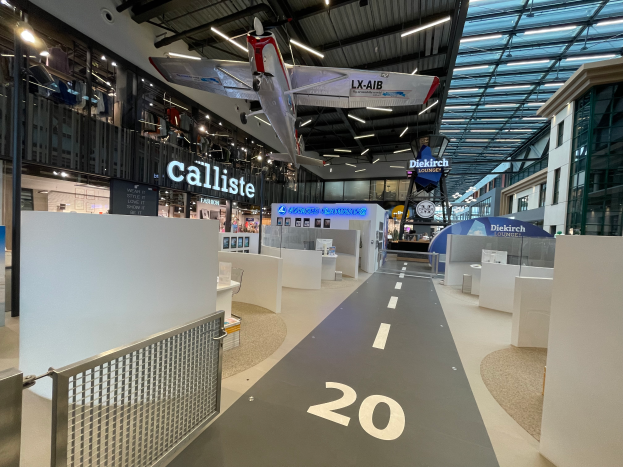 Ein geräumiger Raum mit weißen Wänden, einem aufgehängten Flugzeug, einem Geländer auf der linken Seite, verstreuten Gegenständen auf der rechten Seite, Texttafeln im Hintergrund und an Metallstangen befestigten Deckenleuchten, wahrscheinlich am Düsseldorf Airport.