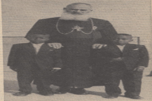 Ein Schwarz-Weiß-Foto eines Mannes, der neben zwei Jungen in Blazern und Krawatten steht, mit Text am unteren Rand, der darauf hindeutet, dass er ihr Vater ist.