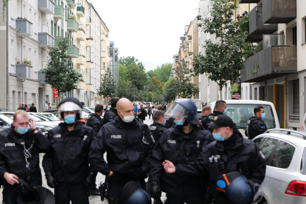 Polizeibeamte in Masken und Helmen gehen eine Straße mit parkenden Autos entlang, flankiert von Gebäuden mit Balkonen und Geländern, unter einem klaren Himmel mit Bäumen im Hintergrund.