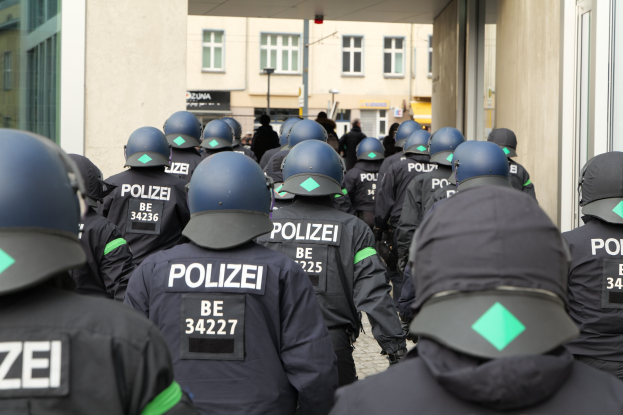 Eine Gruppe von Polizisten in Uniform und Helmen vor einem Gebäude stehend, mit einigen Menschen, Pfählen und anderen Gebäuden im Hintergrund.