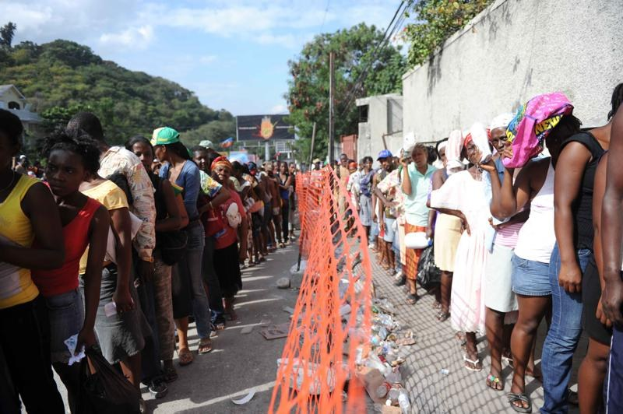Lange Schlange von Menschen, die an einer Wahllokalwarteschlange stehen, mit einem Zaun, einer Wand, Bäumen, Gebäuden, Strommasten, Drähten und einer Werbetafel im Hintergrund.