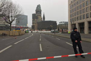 Ein Polizist in schwarzer Uniform und Mütze steht in einer Berliner Straße umgeben von Verkehrskegeln, Fahrrädern, Laternenpfählen, Bäumen und Gebäuden, mit einem Kirchturm und blauem Himmel im Hintergrund.