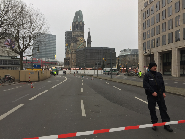 Ein Polizist in schwarzer Uniform und Mütze steht in einer Berliner Straße umgeben von Verkehrskegeln, Fahrrädern, Laternenpfählen, Bäumen und Gebäuden, mit einem Kirchturm und blauem Himmel im Hintergrund.