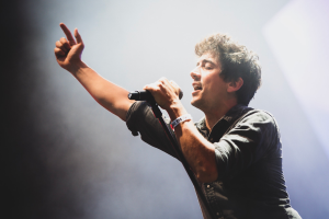 Ein Mann in schwarzem Hemd und Jeans singt in ein Mikrofon auf einer hell erleuchteten Bühne, die Augen geschlossen, während das Publikum im Hintergrund jubelt.