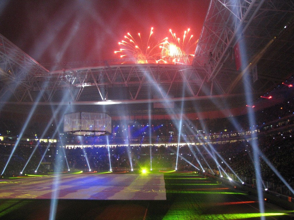 Olympiastadion in London bei Nacht, hell erleuchtet mit grünem Rasen, umgeben von einer großen Menge und Feuerwerk, das den Himmel erhellt.