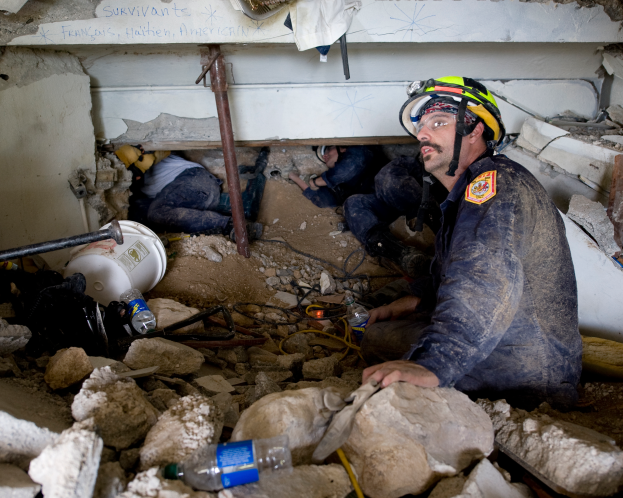 Ein Feuerwehrmann in einem Helm sitzt zwischen Trümmern, darunter Steine und Flaschen, mit einer Wand im Hintergrund, auf der ein Text zu erkennen ist, der den Ort eines israelischen Luftangriffs anzeigt.