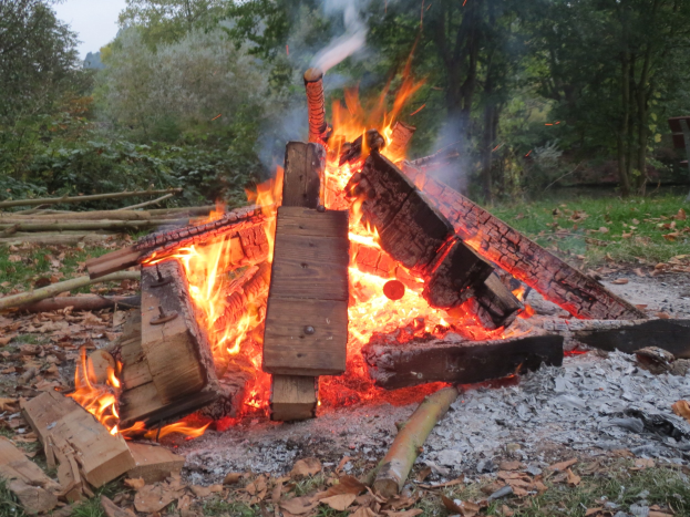 Lagerfeuer in einem bewaldeten Gebiet umgeben von Holzscheiten, Steinen und trockenen Blättern mit Bäumen und einem klaren blauen Himmel im Hintergrund.
