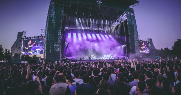 Eine große Menschenmenge bei einem Musikfestival schaut auf die Bühne mit Musikern und Bildschirmen, die vor einem Gebäude, Bäumen und einem klaren blauen Himmel aufgebaut ist.
