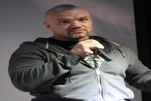 Ein Mann in einer schwarzen Kapuzenjacke und einem schwarzen Anzug mit einem weißen Hemd hält ein Mikrofon und steht selbstbewusst vor einer Wand, er scheint ein Komiker mit zurückgekämmtem Haar zu sein.