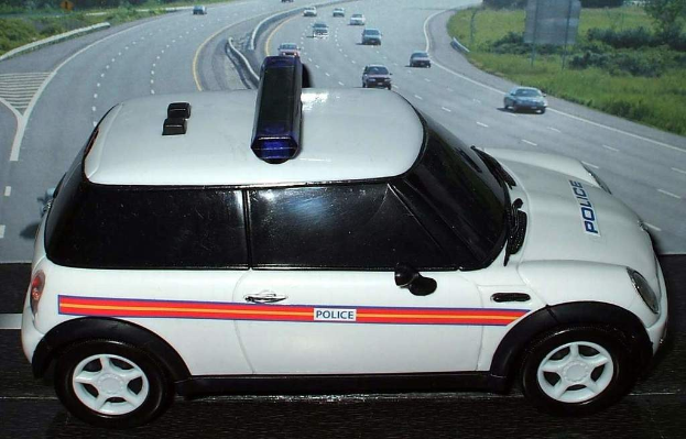Weißer Polizeiwagen mit "Polizei"-Aufschrift auf einer Straße außerhalb einer Stadt, umgeben von bewegendem Verkehr, Gras und Bäumen auf beiden Seiten.