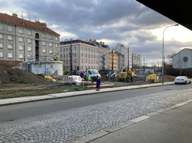 Eine Straße in der Stadt mit parkenden Autos, Gebäuden, Laternen, Bäumen und einem bewölktem Himmel, mit einer Absperrung mit Fahrzeugen und Fussgängern im Vordergrund.