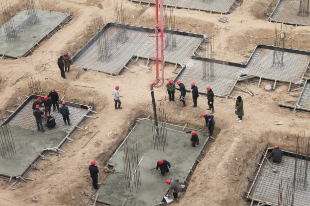 Gruppe von Bauarbeitern mit Helmen auf einer Baustelle, einige halten Werkzeuge und gießen Beton, mit Eisenstangen und einem Pfahl sichtbar.