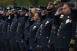 Eine Gruppe von Polizeibeamten in Uniform und Mützen, die in einer Reihe salutieren, vor einem unscharfen Baumhintergrund.