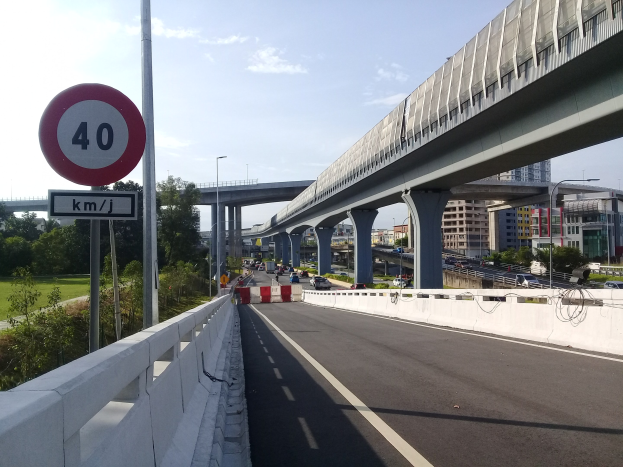 Straße mit Geschwindigkeitsbegrenzungsschild, Fahrzeuge, Brücke mit Pfeilern, Laternen, Bäume, Gebäude und bewölkter Himmel im Hintergrund.