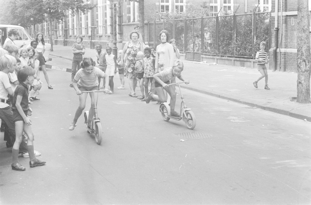 Schwarzes Bild von Kindern, die auf Rollern die Straße entlangfahren, mit einem geparkten Auto auf der linken Seite, Gebäuden, Bäumen, Pfählen und einem Zaun im Hintergrund.