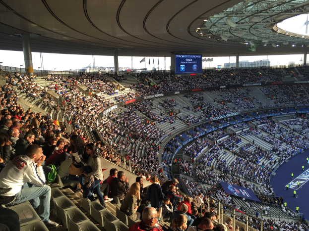 Großes Publikum in einem Stadion bei einem Fußballspiel mit einer Bühne rechts, Fahnen, Stangen, einem Bildschirm und dem Allianz Stadion in München, Deutschland im Hintergrund.