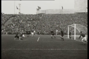 Ein Schwarz-Weiß-Foto von einem WM-Finale 1958 zwischen Manchester United und Liverpool, mit Spielern auf dem Feld, einem Torpfosten rechts, Zuschauern in den Rängen und Fahnen im Hintergrund.