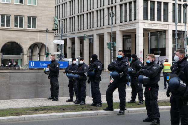 Eine Gruppe von Polizisten in schwarzen Uniformen und Masken steht vor einem Gebäude mit Glasfenstern und Säulen in Berlin, Deutschland, einige halten Helme, mit Laternenmasten, Verkehrsampeln, Texttafeln, einer Statue und anderen Menschen im Hintergrund.