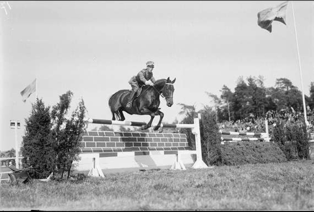Schwarzes und weiße Foto eines Pferdes und Reiters, die über eine Ziegelwand-Sühranke springen, wobei der Reiter einen Helm trägt und die Szenerie von Pflanzen, Flaggen, Gras, Bäumen und einem klaren Himmel umgeben ist.