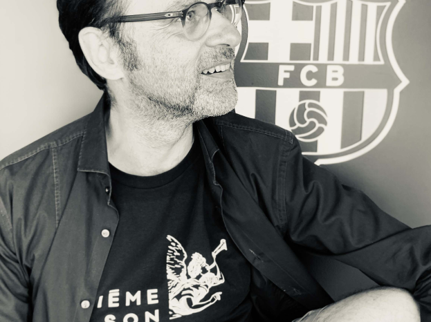Ein Mann mit Brille und schwarzem Hemd sitzt lachend vor einem schwarz-weißen FC Barcelona-Logo.