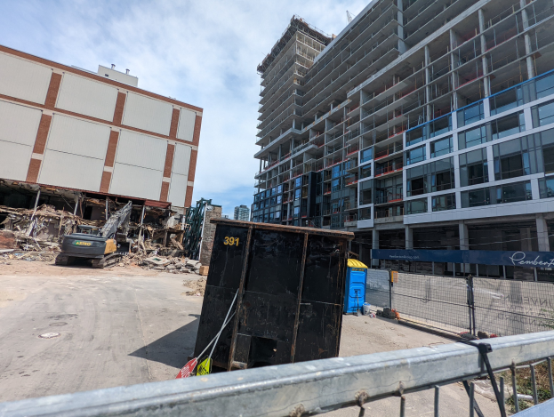 Großes Gebäude mit Schutt davor, umgeben von einem Metallzaun, einem Container, Pflanzen, Bäumen, Pfählen, einem Schild, Fahrzeugen, Steinen und einem bewölkten Himmel, was auf eine Baustelle für einen neuen Wohnkomplex hinweist.