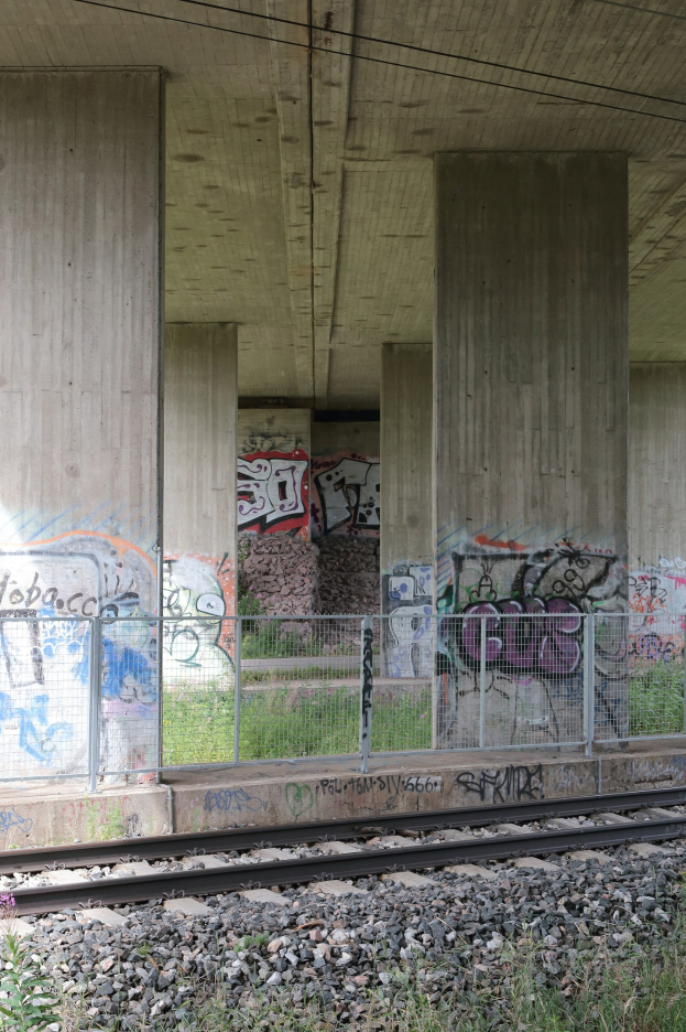 Eisenbahnschiene mit Graffiti an einer Seitenstruktur, umgeben von Steinen, Gras, einem Zaun, Pfeilern, Überlandleitungen und einem Dach.