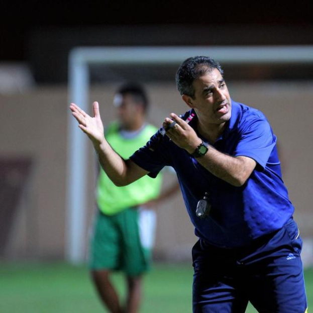 Ein Mann in einem blauen T-Shirt und Hose steht auf einem Fußballfeld und hält ein Mikrofon in der Hand, während er Spielern zunickt. Im Hintergrund sind eine Person in einem grünen Kleid, ein Torpfosten und eine Wand zu sehen.