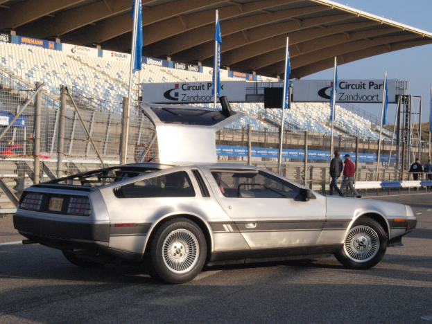 Ein silberner DeLorean DMC-12 Wagen auf einer Straße umgeben von Menschen, Flaggen, Schildern, Geländern, Stühlen und einer Hütte, mit Bäumen und einem klaren blauen Himmel im Hintergrund.