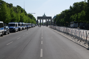 Eine Reihe von Polizeiwagen, die auf einer Straße vor dem Brandenburger Tor in Berlin, Deutschland, geparkt sind, mit Menschen auf Fahrrädern und auf der Straße stehend, Barrieren, Bäume und ein Tor mit Statuen im Hintergrund.