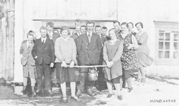 Schwarzes und weißes Foto von Menschen, die lächeln und eine Schleife vor einem Schulgebäude mit einem Fenster und einer Tür halten.