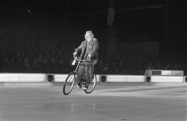 Schwarzes und weißes Bild eines Mannes, der auf einem Eisstadion Fahrrad fährt, mit Zuschauern im Hintergrund und einer Wand dahinter.