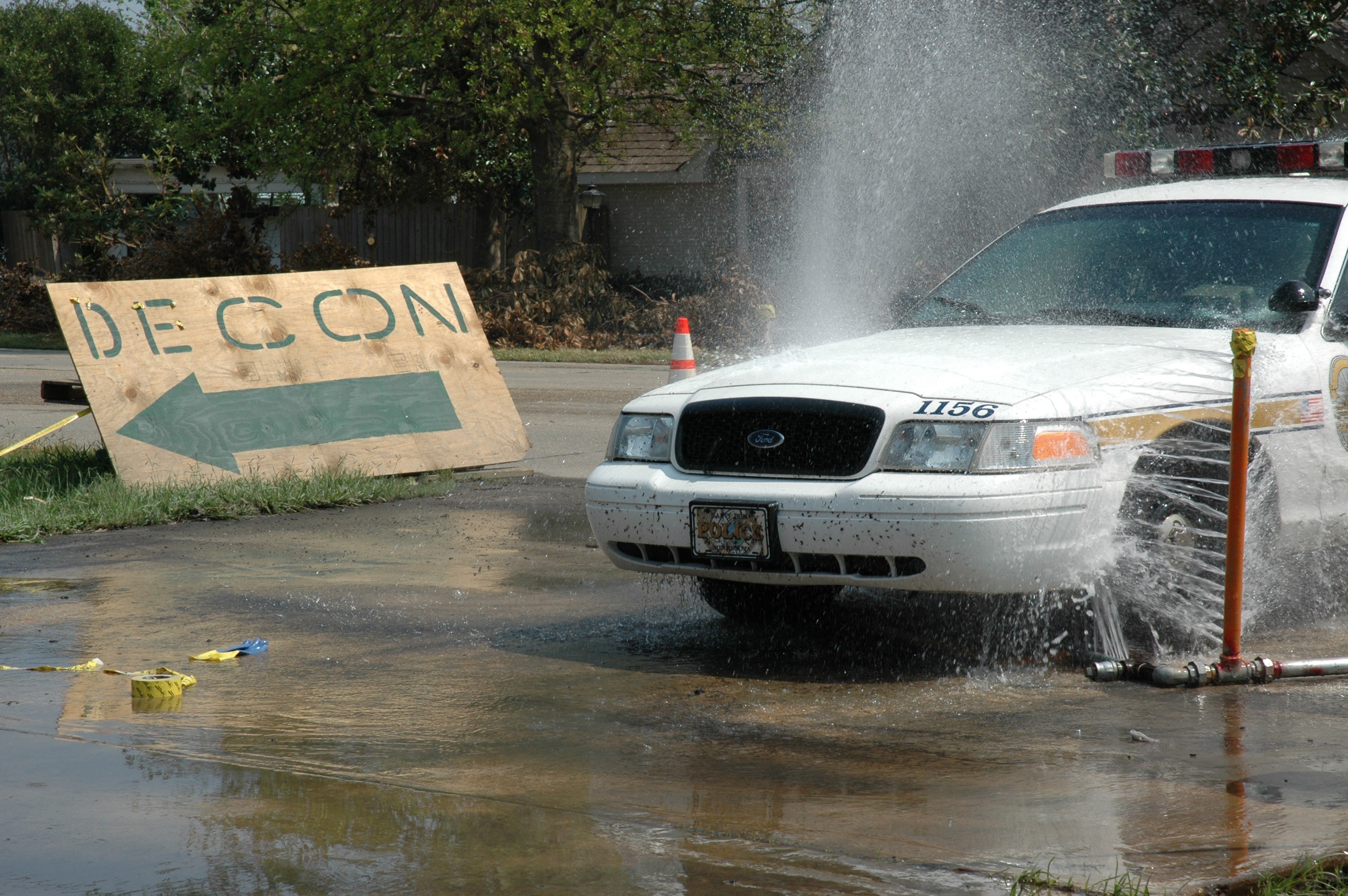 Ein Streifenwagen fährt durch eine Pfütze, mit einem Schild, einer Verkehrskegel und anderen Gegenständen am Boden, mit Bäumen, Gebäuden und einem klaren blauen Himmel im Hintergrund.